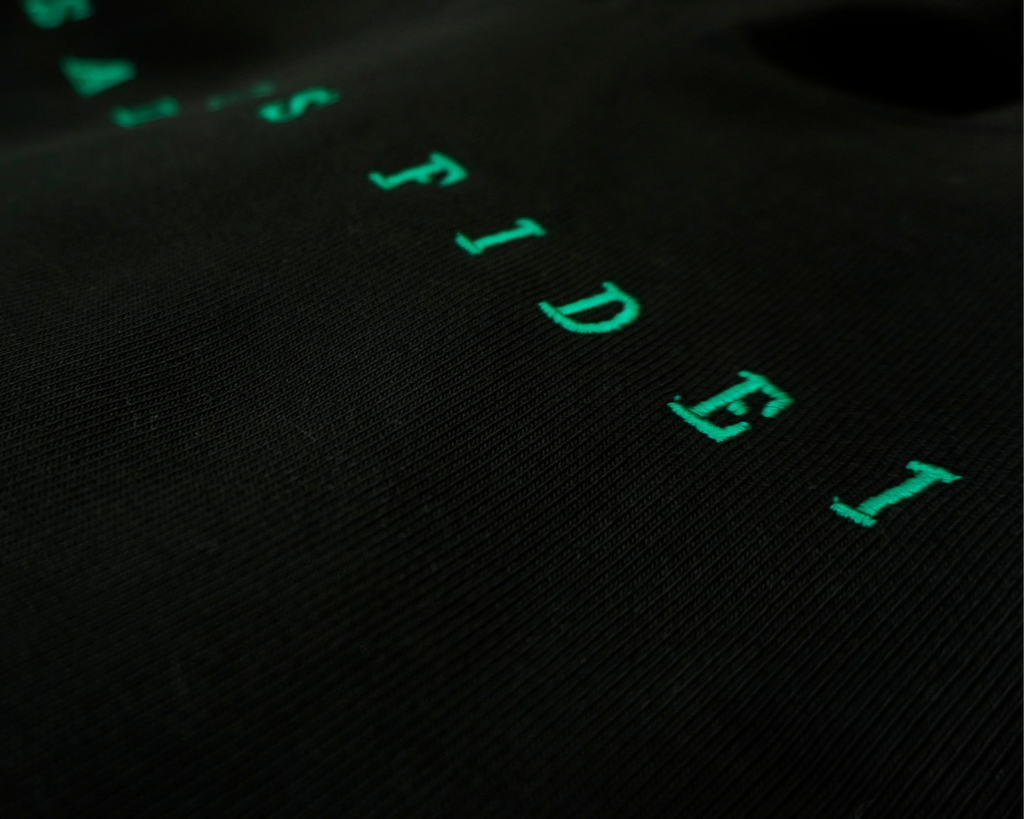 Close-up of a black textured fabric surface with bright green embroidered or printed letters spelling part of a word, photographed in low light.