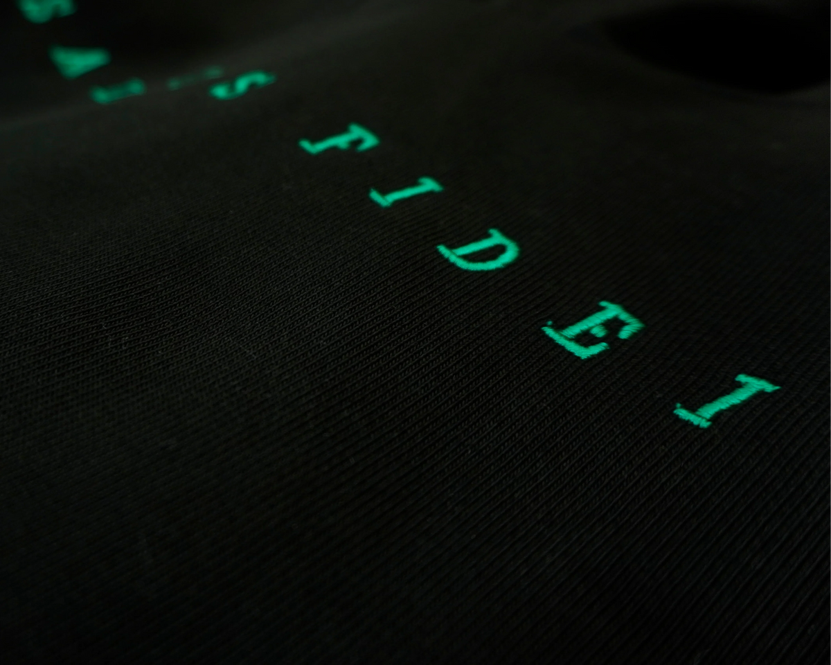 Close-up of a black textured fabric surface with bright green embroidered or printed letters spelling part of a word, photographed in low light.