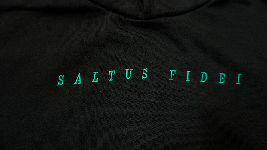 Close-up of a black hoodie featuring the phrase 'SALTUS FIDEI' printed across the chest in a bright green serif font.